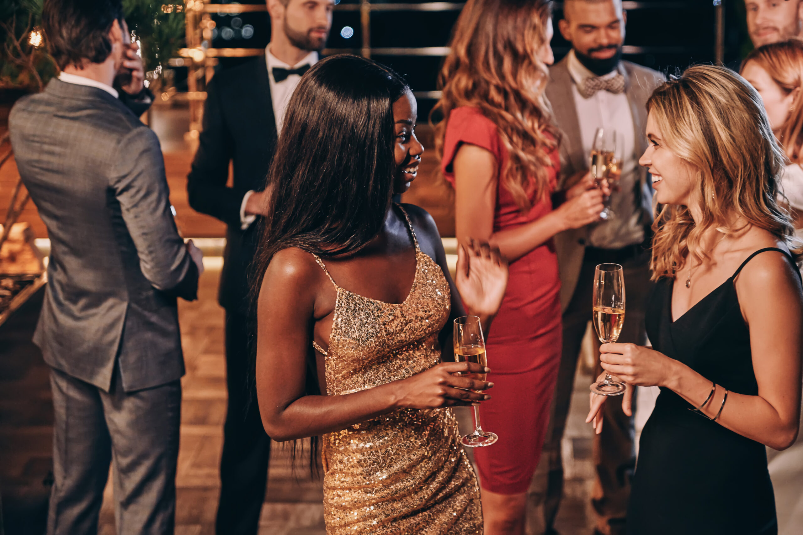 Group of beautiful young people in formalwear communicating and A group of well-dressed people socializing at an elegant evening event, with two women in the foreground holding champagne glasses and smiling at each other.