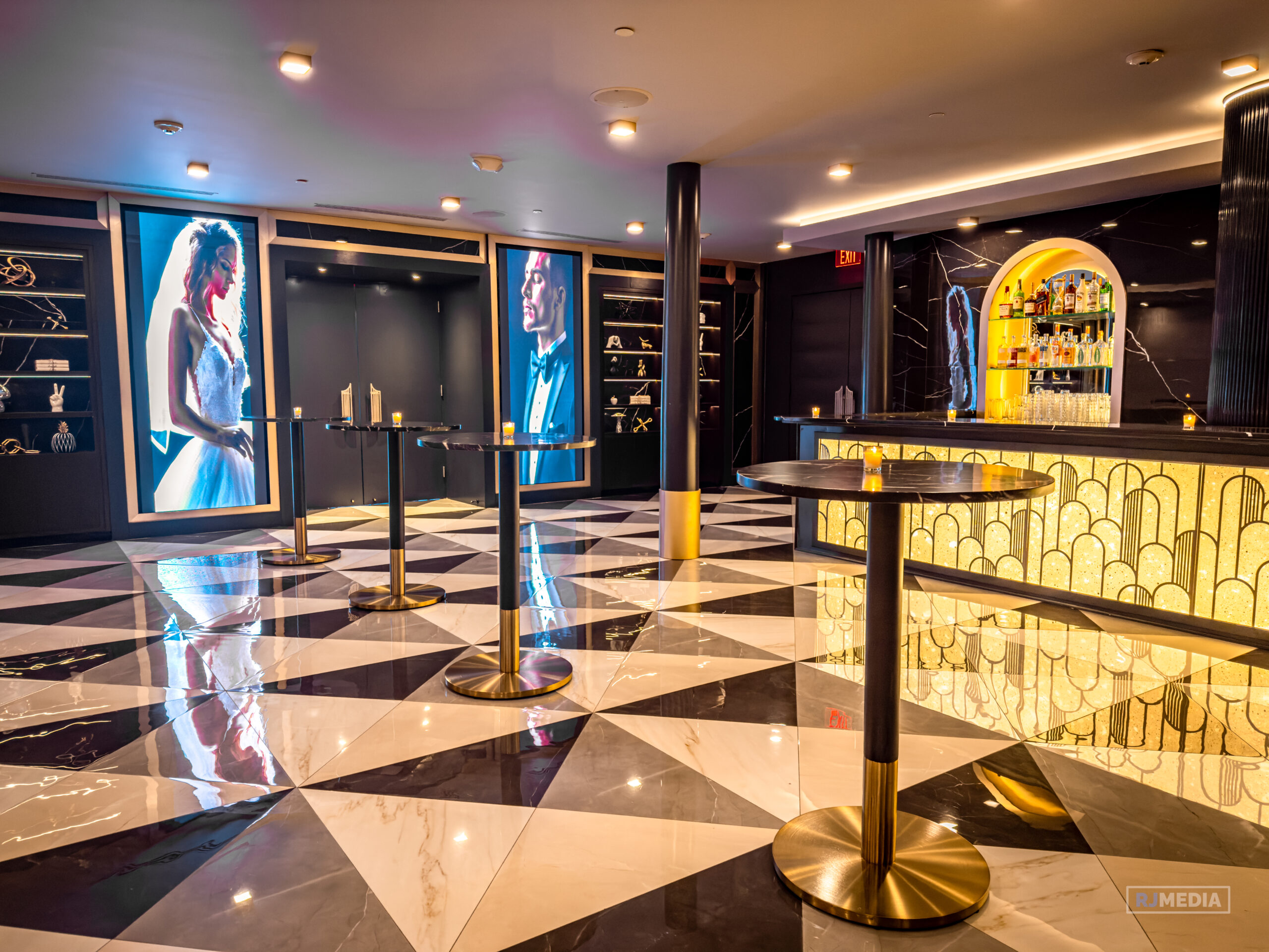 Modern cocktail lounge with black-and-white geometric tile floor, high-top tables, a gold-lit bar with bottles, and large illuminated portraits on the wall.
